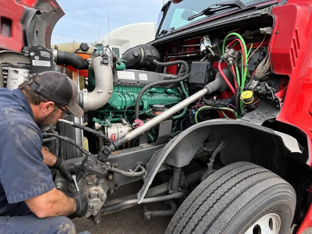 Diesel technician servicing the frame-mounted power steering gear and steering linkage on a heavy-duty commercial truck with the engine bay open.