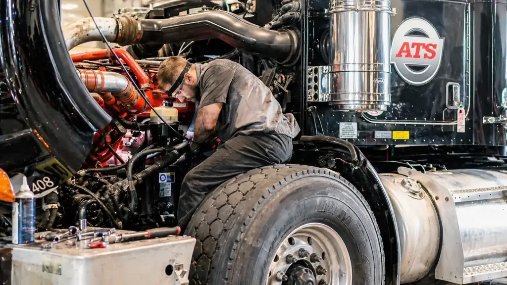 diesel mechanic using a heavy duty transmission jack to remove a semi truck transmission in a commercial repair shop showing drivetrain service tools and clutch housing