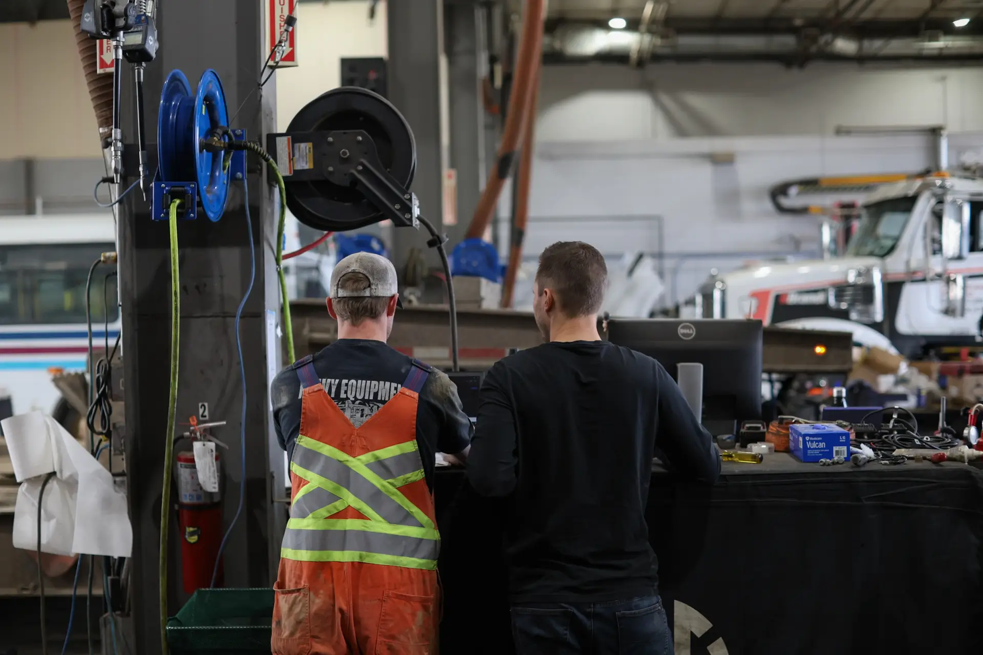 Service manager using digital shop management software on a tablet while diesel technicians perform repairs on a Class 8 semi truck inside a modern heavy-duty repair facility.